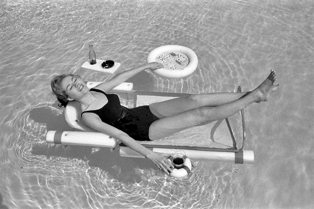 Sunbathing with the latest swimming pool equipment, including a lounge chair with floating beverage holders and game table attachments.. 1961