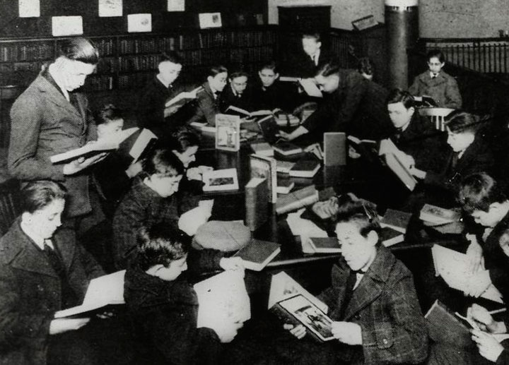 Work with schools : after a book talk, showing boys gathered around table gathering. New York, 1920