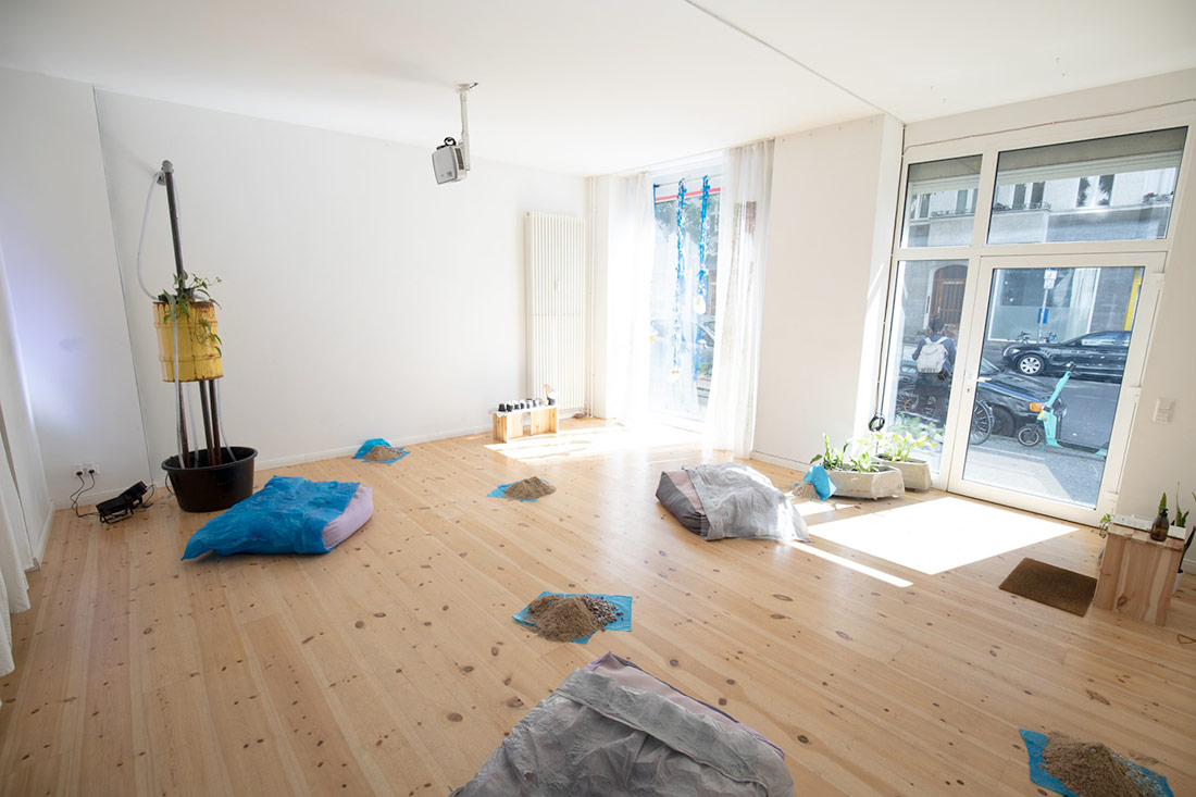An exhibition view of “Conspiring Timelines, Shimmering Temporalities” is shown inside the exhibition space, TIER (Berlín). Across the warm wooden floor are pillows covered in fused plastic pillowcases, piles of marbles and sand and a projection of a video showing elderflower leaves being cut together and apart from trees.